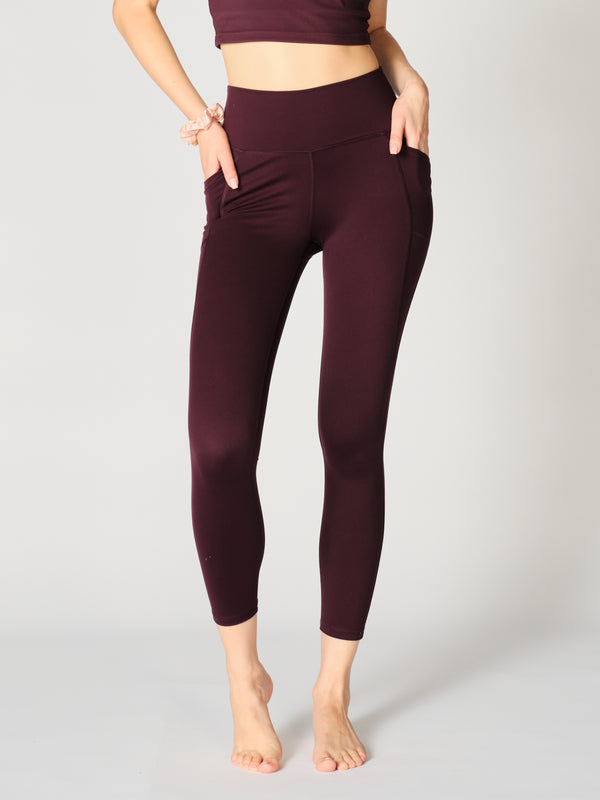 Close up view of X by Gottex Rachel ankle legging in color Merlot, that accentuate the contours of the model’s legs. An image shows the two side pockets that can fit a phone, a credit card, a set of keys, or other personal items. The design appears to be high-waisted, ankle length, tummy-control flat wide waistband, strategic placement of the seams flatter curves and elongate legs. The fabric appears to be soft, 4-way stretch, with some compression. The legging is well-suited for yoga, pilates, barre, gym.