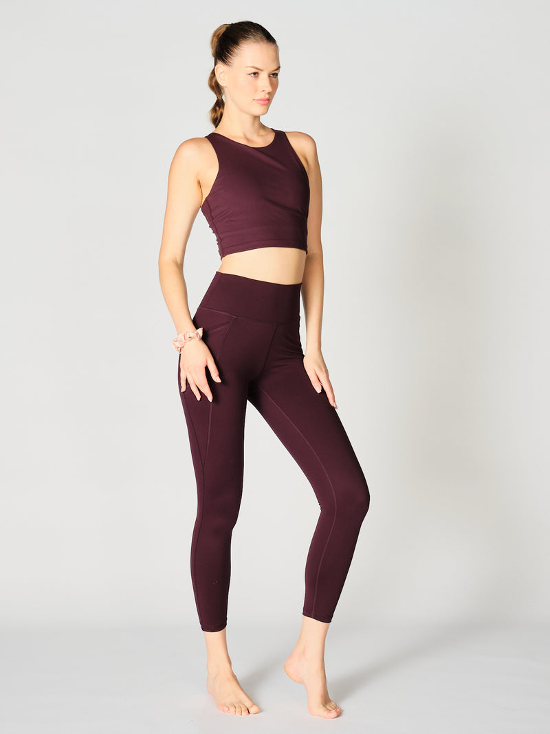 A model wears the X by Gottex Rachel Ankle Legging in a sophisticated merlot shade. She pairs the leggings with a coordinating crop top that complements the high-waisted cut, highlighting a sleek and modern athletic silhouette. The leggings feature contouring seams that run down the legs, suggesting a design that both flatters and supports. The fabric appears to be soft, 4-way stretch, with some compression. The legging is well-suited for yoga, pilates, barre, gym, and an active lifestyle in general