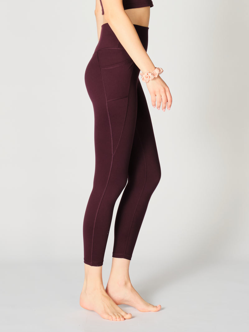 Close up of the right side of the X by Gottex Rachel ankle legging in color Merlot. The legging contours of the model’s legs, it has a side pocket that can fit a phone, a credit card, a set of keys, or other personal items. The design appears to be high-waisted, ankle length, tummy-control flat wide waistband, strategic placement of the seams flatter curves and elongate legs. The fabric appears to be soft, 4-way stretch, with some compression. The legging is well-suited for yoga, pilates, barre, gym.