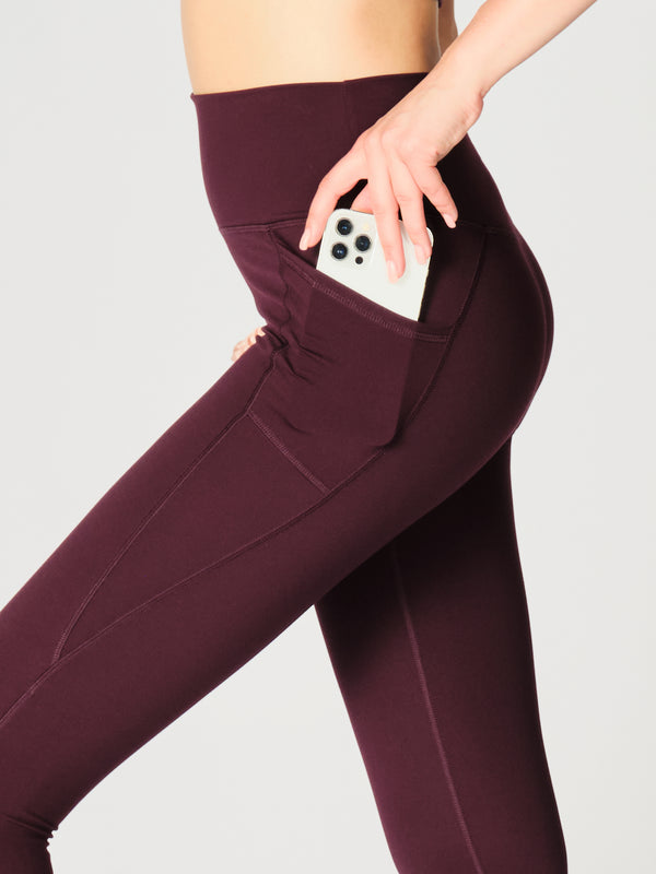 A close-up of a X by Gottex Rachel Ankle Leggings in Merlot. The leggings have a high-rise waist, providing a secure and flattering fit. On the left side, there's a conveniently placed pocket with a smartphone halfway inserted. The fabric of the leggings appears smooth and stretchy. The merlot hue is deep and vivid, giving the leggings a versatile and stylish appeal. The legging is well-suited for yoga, pilates, barre, gym, and an active lifestyle in general.