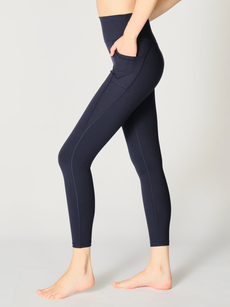 Close up view of the left side of the X by Gottex Rachel ankle legging in Midnight. The legging contours of the model’s legs, it has a side pocket that can fit a phone, a credit card, a set of keys, or other personal items. The design appears to be high-waisted, ankle length, tummy-control flat wide waistband, strategic placement of the seams flatter curves and elongate legs. The fabric appears to be soft, 4-way stretch, with some compression. The legging is well-suited for yoga, pilates, barre, gym