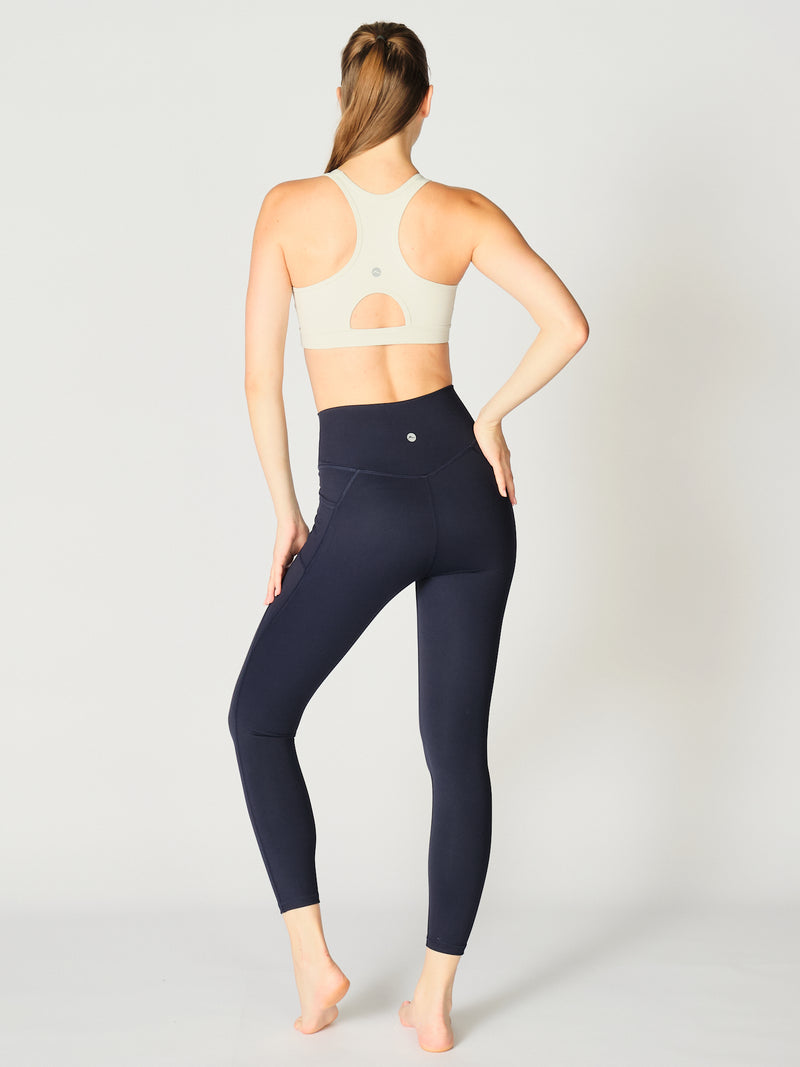 A model is posed with her back to the camera, turned to look over her shoulder, showcasing the X by Gottex Rachel Ankle Legging in Midnight. She’s also wearing a matching sports bra with an active racer back cut. The design appears to be high-waisted, ankle length, tummy-control flat wide waistband, strategic placement of the seams flatter curves and elongate legs. The fabric appears to be soft, 4-way stretch, with some compression. The legging is well-suited for yoga, pilates, barre, gym.