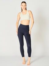 A model wears the X by Gottex Rachel Ankle Legging in a sophisticated Midnight shade. She pairs the leggings with a pearl sports bra that complements the high-waisted cut, highlighting a sleek and modern athletic silhouette. The leggings feature contouring seams that run down the legs, suggesting a design that both flatters and supports. The fabric appears to be soft, 4-way stretch, with some compression. The legging is well-suited for yoga, pilates, barre, gym, and an active lifestyle in general.  